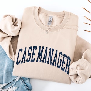 May include: A beige sweatshirt with the text "CASE MANAGER" in navy blue arched across the front. A silver necklace with a small pendant is on the sweatshirt. The sweatshirt is folded with the sleeves tucked in. Blue jeans are visible.