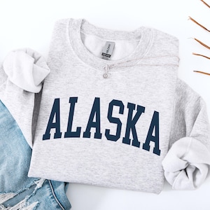 May include: A light gray sweatshirt with the word "ALASKA" in navy blue arched lettering. The sweatshirt is folded with the sleeves rolled up. A pair of blue jeans is visible on the left side of the image.