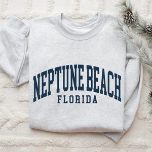 May include: A light gray sweatshirt with the words "NEPTUNE BEACH FLORIDA" in navy blue arched lettering. The sweatshirt is folded on a white textured surface, with pine branches and pinecones in the background.