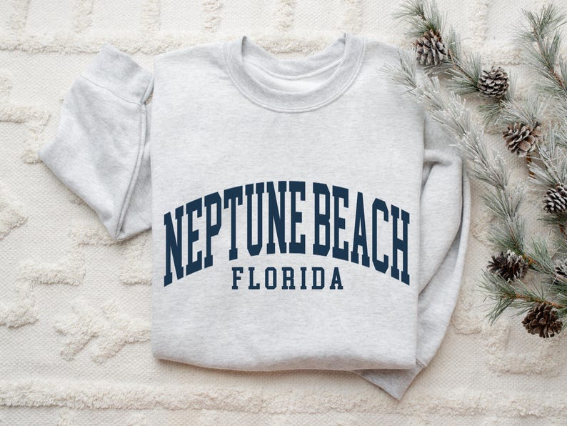 May include: A light gray sweatshirt with the words "NEPTUNE BEACH FLORIDA" in navy blue arched lettering. The sweatshirt is folded on a white textured surface, with pine branches and pinecones in the background.