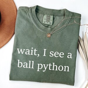 K&ouml;nnte beinhalten: Ein olivgr&uuml;nes Kurzarm-T-Shirt mit Rundhalsausschnitt. Auf der Vorderseite ist der wei&szlig;e Schriftzug "wait, I see a ball python" aufgedruckt. Ein kleines Etikett im Kragen zeigt "Comfort Colors" und die Gr&ouml;&szlig;enangabe "S-P-CH".