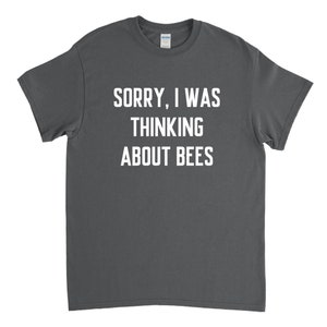 以下が含まれることがあります： 白い文字で「SORRY, I WAS THINKING ABOUT BEES」とプリントされた濃いグレーのTシャツです。