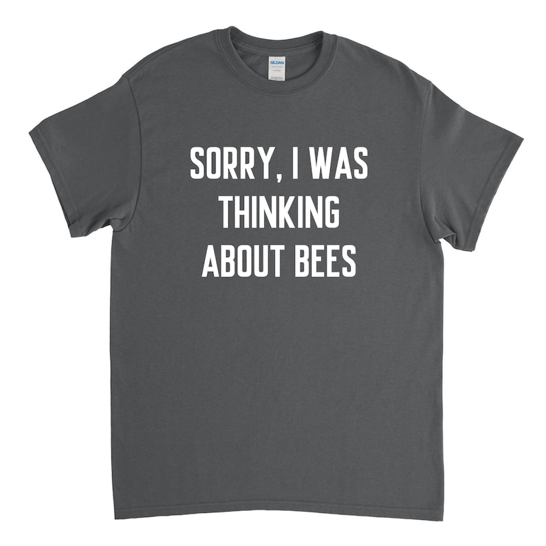 以下が含まれることがあります： 白い文字で「SORRY, I WAS THINKING ABOUT BEES」とプリントされた濃いグレーのTシャツです。