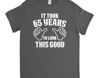 May include: Dark gray t-shirt with white text that reads "It took 65 years to look this good" with two white thumbs up symbols.