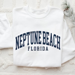 May include: A white sweatshirt with the text "NEPTUNE BEACH FLORIDA" in navy blue arched lettering. The sweatshirt is folded on a white textured surface, with a sprig of greenery and pinecones in the background.