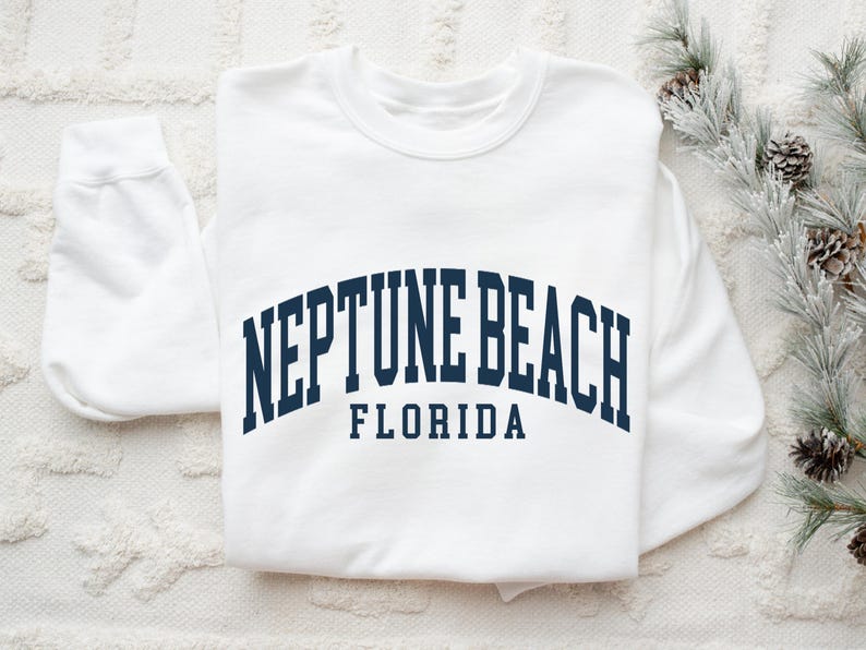 May include: A white sweatshirt with the text "NEPTUNE BEACH FLORIDA" in navy blue arched lettering. The sweatshirt is folded on a white textured surface, with a sprig of greenery and pinecones in the background.