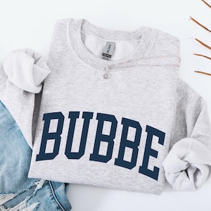 May include: A light gray sweatshirt with the word "BUBBE" in navy blue block letters. The sweatshirt is folded with the sleeves visible, and is next to a pair of blue jeans.