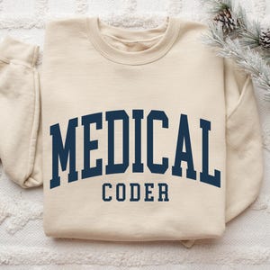 May include: A cream-colored sweatshirt with the words "MEDICAL CODER" in navy blue block letters. The sweatshirt is a crew neck style and has long sleeves. The image is taken from a top-down perspective.
