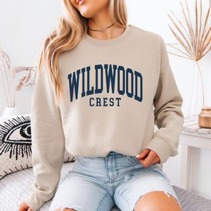 May include: A beige sweatshirt with the text "WILDWOOD CREST" in navy blue arched lettering. The sweatshirt is a crew neck style and appears to be made of a soft material. The model is wearing light blue denim shorts.