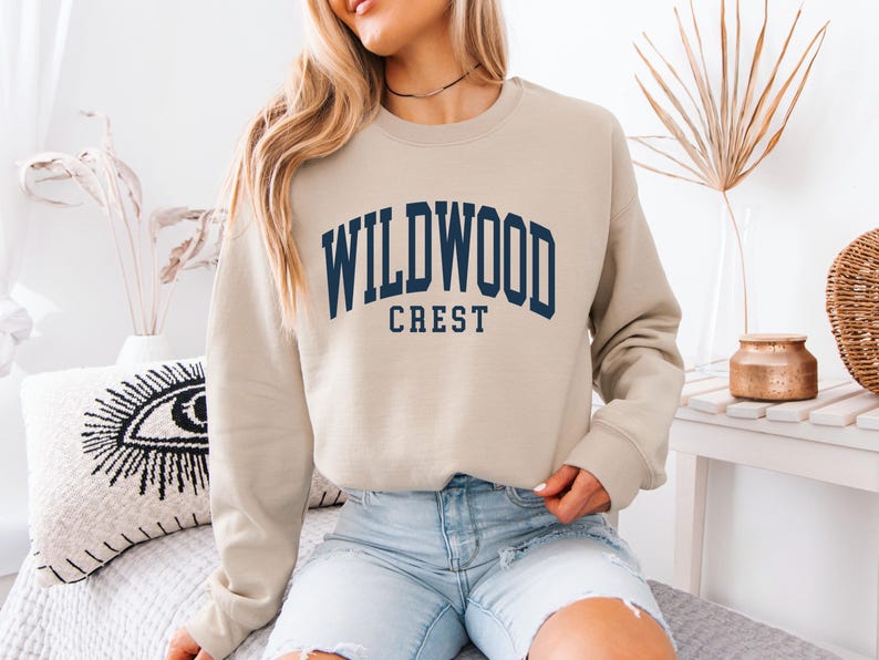 May include: A beige sweatshirt with the text "WILDWOOD CREST" in navy blue arched lettering. The sweatshirt is a crew neck style and appears to be made of a soft material. The model is wearing light blue denim shorts.