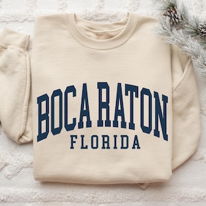 May include: Cream-colored sweatshirt with the text "BOCA RATON FLORIDA" in navy blue block letters. The sweatshirt is a crew neck style and has long sleeves. The image is taken from a top-down perspective.