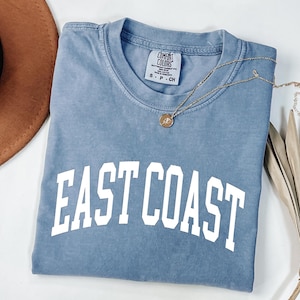 May include: A blue short-sleeve t-shirt with the words "EAST COAST" in white block letters. A gold necklace with a pendant rests on the shirt. A brown hat and dried plant are in the background.