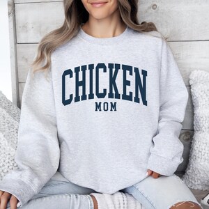 May include: A light gray sweatshirt with the text "CHICKEN MOM" in navy blue block letters. The word "MOM" is centered below "CHICKEN". The sweatshirt is a crew neck style and appears to be made of a soft material.