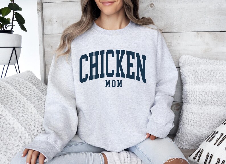 May include: A light gray sweatshirt with the text "CHICKEN MOM" in navy blue block letters. The word "MOM" is centered below "CHICKEN". The sweatshirt is a crew neck style and appears to be made of a soft material.