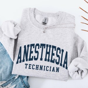 May include: A light gray sweatshirt with the words "ANESTHESIA TECHNICIAN" in navy blue arched lettering. The sweatshirt is folded, with the sleeves visible. A silver necklace with a small pendant is on the sweatshirt. Blue jeans are visible on the left.