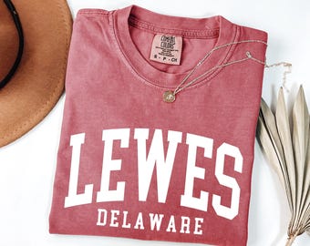 Puede incluir: Una camiseta de manga corta de color rosa polvoriento con el texto blanco LEWES DELAWARE en una fuente universitaria en negrita. La camiseta tiene un cuello redondo y una etiqueta de la marca Comfort Colors en el interior con la talla S.