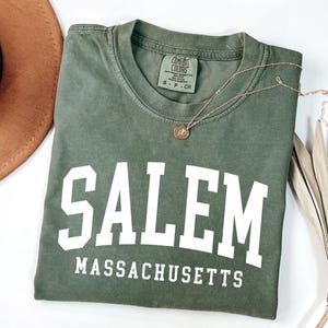 Puede incluir: Camiseta verde oliva con la palabra "SALEM" en letras blancas grandes y "MASSACHUSETTS" debajo. Un collar dorado con un colgante descansa sobre la camiseta. Un sombrero marr&oacute;n y una planta seca est&aacute;n en el fondo.