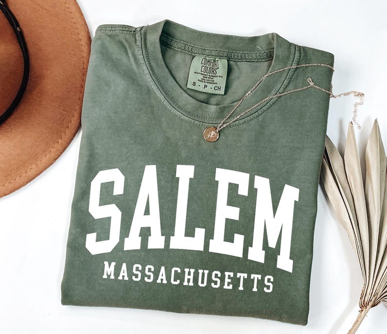 Puede incluir: Camiseta verde oliva con la palabra "SALEM" en letras blancas grandes y "MASSACHUSETTS" debajo. Un collar dorado con un colgante descansa sobre la camiseta. Un sombrero marr&oacute;n y una planta seca est&aacute;n en el fondo.
