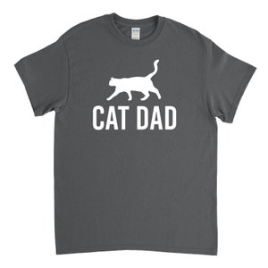 Puede incluir: Una camiseta gris oscuro con una silueta blanca de un gato caminando y el texto "CAT DAD" impreso en la parte delantera.