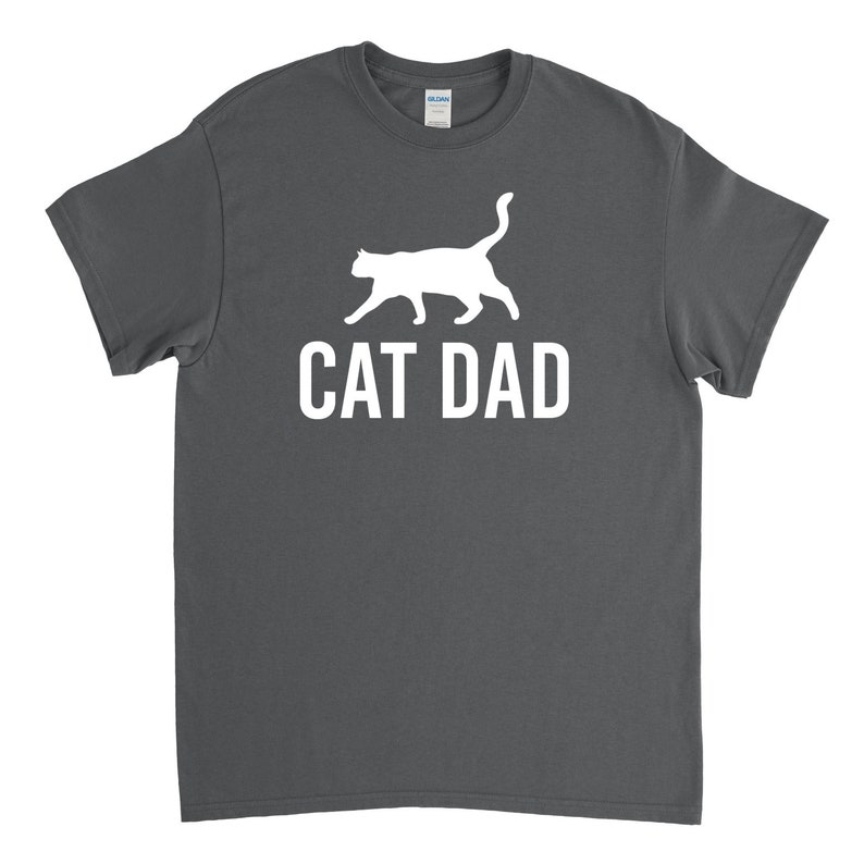 Puede incluir: Una camiseta gris oscuro con una silueta blanca de un gato caminando y el texto "CAT DAD" impreso en la parte delantera.