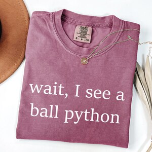 K&ouml;nnte beinhalten: Ein malvenfarbenes Kurzarm-T-Shirt mit Rundhalsausschnitt und dem wei&szlig;en Schriftzug "wait, I see a ball python" auf der Vorderseite. Das T-Shirt hat ein Comfort Colors-Markenetikett am Innenkragen, das die Gr&ouml;&szlig;e Small angibt.