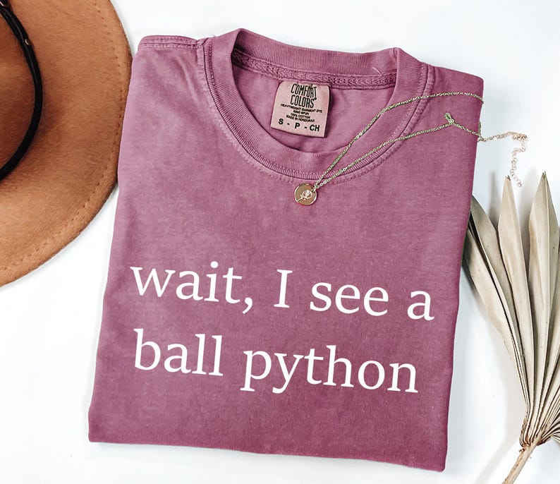 K&ouml;nnte beinhalten: Ein malvenfarbenes Kurzarm-T-Shirt mit Rundhalsausschnitt und dem wei&szlig;en Schriftzug "wait, I see a ball python" auf der Vorderseite. Das T-Shirt hat ein Comfort Colors-Markenetikett am Innenkragen, das die Gr&ouml;&szlig;e Small angibt.
