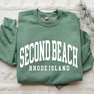 May include: A sage green sweatshirt with the words "SECOND BEACH RHODE ISLAND" printed in white, arched across the chest. The sweatshirt is folded neatly on a white textured surface, with a sprig of faux pine needles and pinecones in the background.