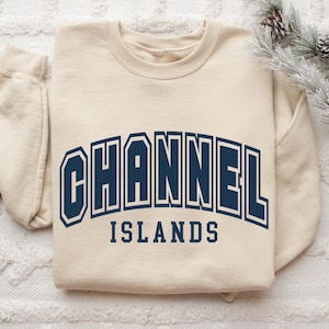Pu&ograve; includere: Felpa color crema con la scritta "CHANNEL ISLANDS" in lettere maiuscole blu navy. Il testo &egrave; arcuato sulla parte anteriore della felpa. La felpa &egrave; appoggiata su una superficie bianca e strutturata.