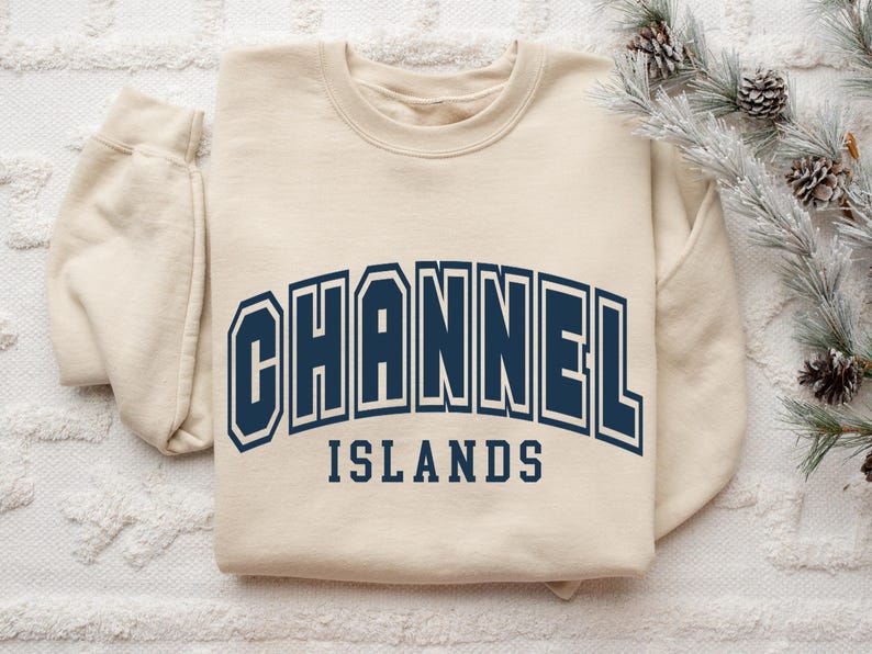 Pu&ograve; includere: Felpa color crema con la scritta "CHANNEL ISLANDS" in lettere maiuscole blu navy. Il testo &egrave; arcuato sulla parte anteriore della felpa. La felpa &egrave; appoggiata su una superficie bianca e strutturata.