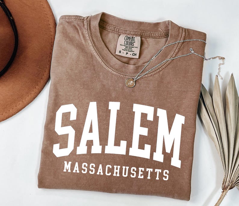 Puede incluir: Una camiseta marr&oacute;n con la palabra "SALEM" en letras blancas grandes, con "MASSACHUSETTS" debajo. Tambi&eacute;n se ve un collar plateado con un colgante y un sombrero marr&oacute;n. La camiseta es de un material suave.