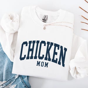 May include: White crewneck sweatshirt with the text "CHICKEN MOM" in navy blue arched lettering. A silver necklace with a small pendant rests on the sweatshirt. The sweatshirt is folded next to a pair of blue jeans.