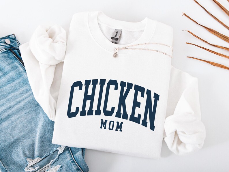 May include: White crewneck sweatshirt with the text "CHICKEN MOM" in navy blue arched lettering. A silver necklace with a small pendant rests on the sweatshirt. The sweatshirt is folded next to a pair of blue jeans.