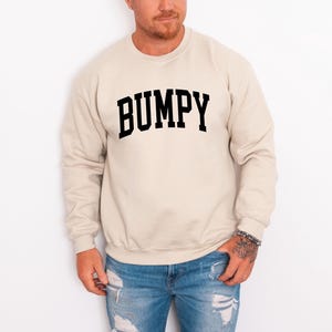 May include: A beige sweatshirt with the word "BUMPY" printed in bold, black, block letters. The sweatshirt is a crew neck style. The person in the image is wearing blue jeans.