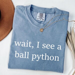 K&ouml;nnte beinhalten: Ein blaues kurz&auml;rmeliges T-Shirt mit Rundhalsausschnitt und dem wei&szlig;en Schriftzug "wait, I see a ball python" auf der Vorderseite. Das T-Shirt hat ein Comfort Colors Markenetikett im Kragen und besteht aus Baumwollstoff.