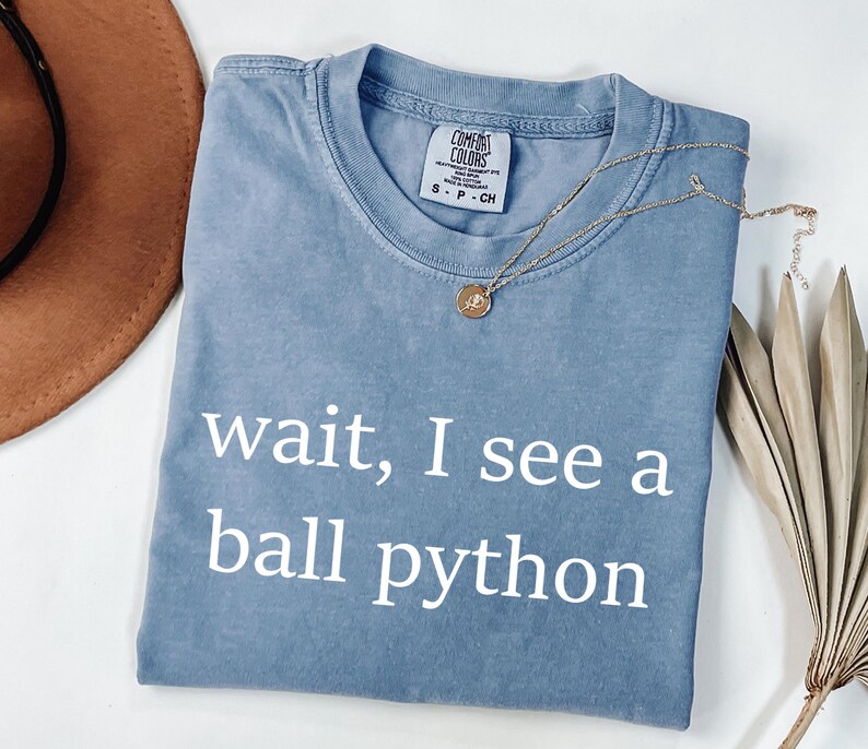 K&ouml;nnte beinhalten: Ein blaues kurz&auml;rmeliges T-Shirt mit Rundhalsausschnitt und dem wei&szlig;en Schriftzug "wait, I see a ball python" auf der Vorderseite. Das T-Shirt hat ein Comfort Colors Markenetikett im Kragen und besteht aus Baumwollstoff.