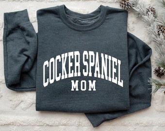 May include: A dark gray sweatshirt with the words "COCKER SPANIEL MOM" in white, arched lettering. The sweatshirt is folded, with the sleeves visible. The design is centered on the chest.
