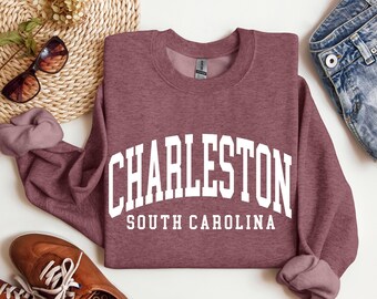 May include: A maroon crewneck sweatshirt with the word "CHARLESTON" in large white block letters arched across the chest, with "SOUTH CAROLINA" below. The sweatshirt is paired with blue denim shorts and brown shoes.