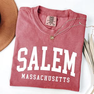 Puede incluir: Una camiseta rosa descolorida con la palabra "SALEM" en letras blancas grandes y "MASSACHUSETTS" debajo. Un collar dorado con un colgante descansa sobre la camiseta. Un sombrero marr&oacute;n y una planta seca est&aacute;n en el fondo.