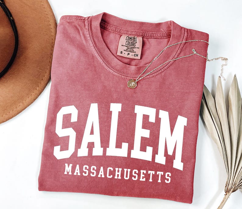 Puede incluir: Una camiseta rosa descolorida con la palabra "SALEM" en letras blancas grandes y "MASSACHUSETTS" debajo. Un collar dorado con un colgante descansa sobre la camiseta. Un sombrero marr&oacute;n y una planta seca est&aacute;n en el fondo.