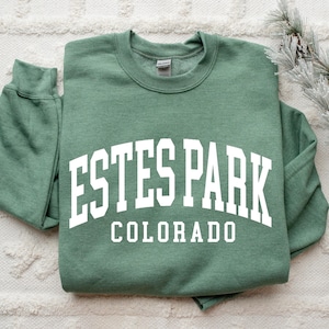 May include: A sage green sweatshirt with the text "ESTES PARK COLORADO" in white, arched across the chest. The sweatshirt is folded and lies on a textured white surface, with a decorative pine branch in the background.