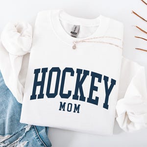 Op de afbeelding: Een witte sweatshirt met de woorden "HOCKEY MOM" in marineblauwe blokletters. De sweatshirt is opgevouwen met een spijkerbroek en een zilveren ketting. De sweatshirt is gemaakt van een zacht materiaal.