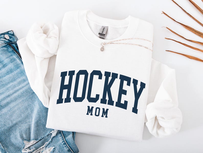 Op de afbeelding: Een witte sweatshirt met de woorden "HOCKEY MOM" in marineblauwe blokletters. De sweatshirt is opgevouwen met een spijkerbroek en een zilveren ketting. De sweatshirt is gemaakt van een zacht materiaal.