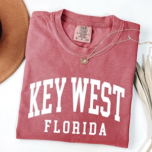 May include: A red t-shirt with the words "KEY WEST FLORIDA" in white block letters. The shirt has a crew neck and short sleeves. A gold necklace with a pendant is draped across the shirt.