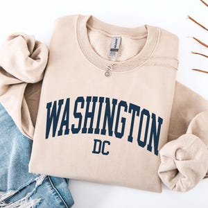 May include: A beige sweatshirt with the word "WASHINGTON" in navy blue arched across the chest, with "DC" below. A silver necklace with a small pendant rests on the sweatshirt. The sweatshirt is folded next to a pair of blue jeans.