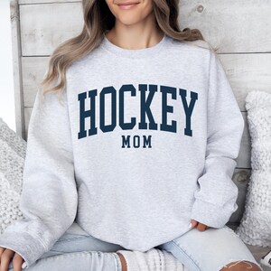 Op de afbeelding: Lichtgrijs sweatshirt met de tekst "HOCKEY MOM" in marineblauwe blokletters. De tekst is in een boog over de borst geplaatst. De sweatshirt heeft een ronde hals en is gemaakt van een zacht materiaal.