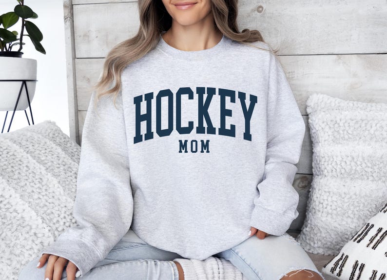 Op de afbeelding: Lichtgrijs sweatshirt met de tekst "HOCKEY MOM" in marineblauwe blokletters. De tekst is in een boog over de borst geplaatst. De sweatshirt heeft een ronde hals en is gemaakt van een zacht materiaal.