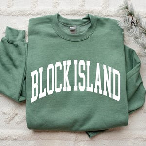May include: A sage green sweatshirt with the words "BLOCK ISLAND" in large, white, arched lettering. The sweatshirt is folded neatly, with the sleeves tucked in. The background is a textured, off-white surface, with a sprig of decorative pine needles and pinecones.