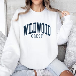 May include: A white sweatshirt with the text "WILDWOOD CREST" in navy blue block letters. The sweatshirt is a crew neck style and appears to be made of a soft material. The image shows the sweatshirt on a person.