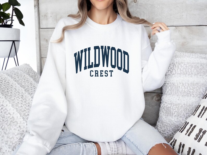 May include: A white sweatshirt with the text "WILDWOOD CREST" in navy blue block letters. The sweatshirt is a crew neck style and appears to be made of a soft material. The image shows the sweatshirt on a person.