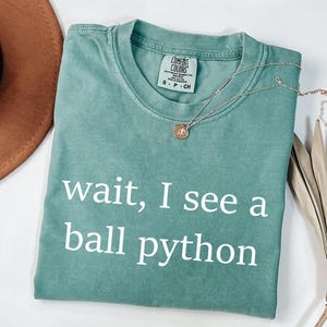 K&ouml;nnte beinhalten: Ein kurz&auml;rmeliges T-Shirt in Salbeigr&uuml;n mit Rundhalsausschnitt und dem wei&szlig;en Text "wait, I see a ball python" auf der Vorderseite. Das Shirt hat ein Comfort Colors Markenetikett am Innenkragen und besteht aus 100% Baumwollstoff.